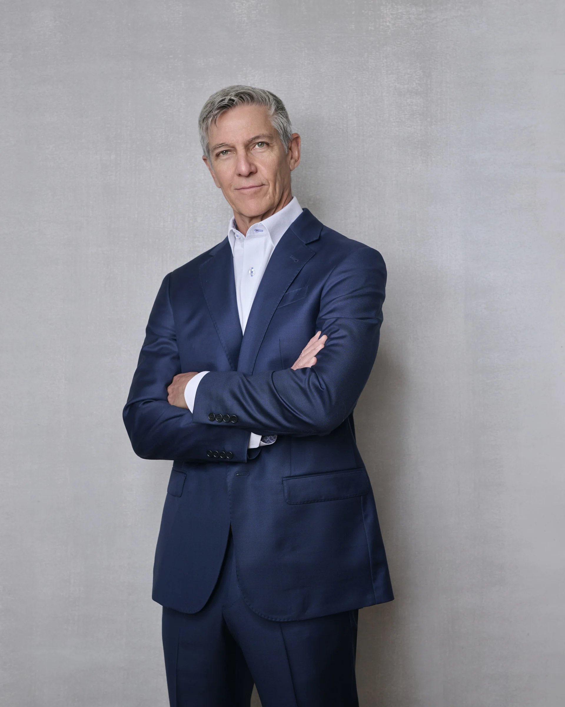 Distinguished executive man in navy blue suit with arms crossed against textured gray backdrop